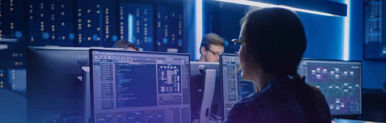 A team of professionals working at computer stations in a darkened room, illuminated by blue and white lights, focused on various screens displaying data and network information.