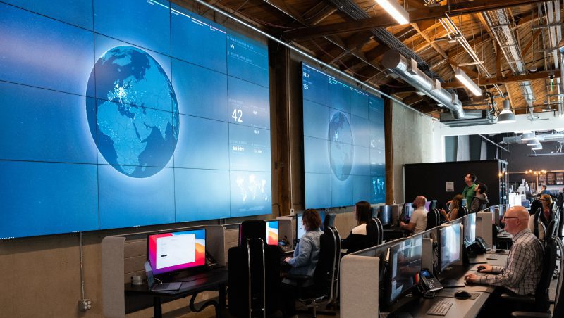 A cybersecurity operations center featuring several workstations with employees monitoring large screens displaying a globe and security metrics, indicating a focus on understanding your security environment and using AI to improve MXDR.