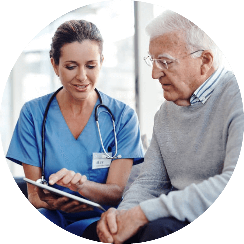 A healthcare professional in a blue uniform discusses information from a tablet with an elderly man wearing glasses and a gray sweater, both seated indoors.