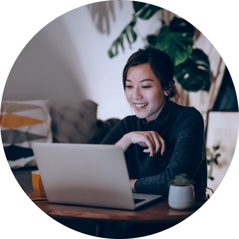 A woman sitting at a wooden table with a laptop in front of her, smiling while working. She is in a cozy room with plants and a couch in the background.
