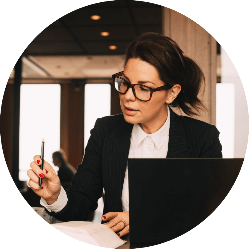 A woman in a pinstriped suit, wearing glasses, is sitting at a table with a laptop. She is holding a pen and appears to be engaged in work, with notes and documents in front of her.