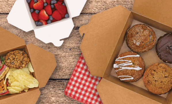 Two brown takeout boxes on a wooden surface. One box contains muffins and the other has a cookie, chips, and a sandwich. A small white box holds assorted berries in the background, next to a red checkered cloth.