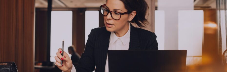 A woman wearing glasses and a blazer is sitting at a table, writing with a pen while looking at a laptop. The environment appears to be a modern office or café.