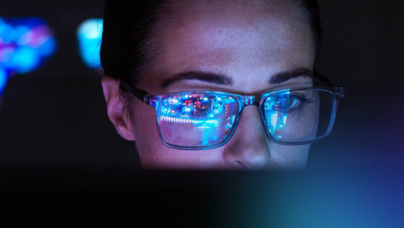 A close-up of a person's face, wearing glasses, with reflections of colorful data visualizations in the lenses, indicating a focus on technology. This image relates to Utilizing Machine Learning Operations and MLOps defined.
