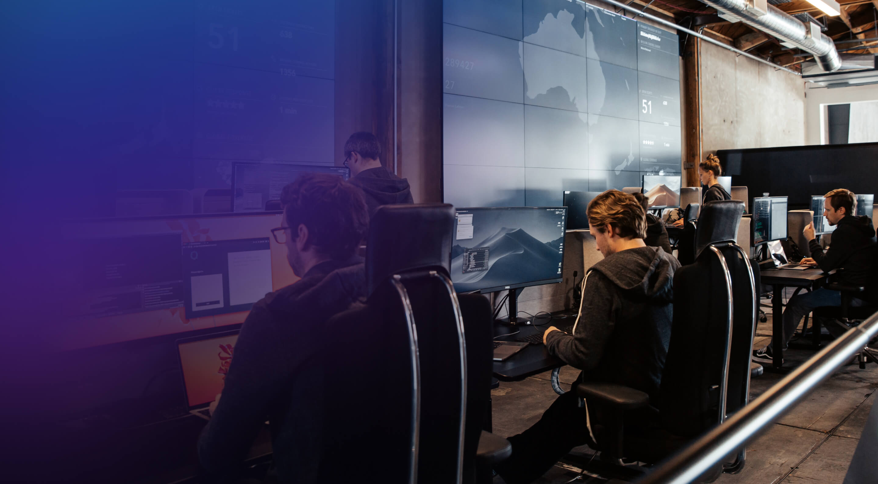 Several people working at computers in a modern office setting. Large screens display data and maps in the background. The environment is bright and organized, highlighting a technology-focused workspace.