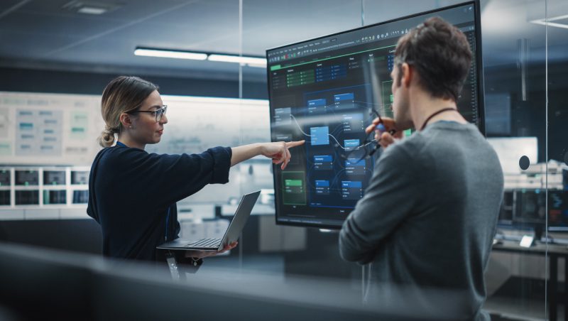A woman holding a laptop points at a large screen displaying data and diagrams, while a man listens attentively. They are in a modern workspace with multiple monitors in the background.
