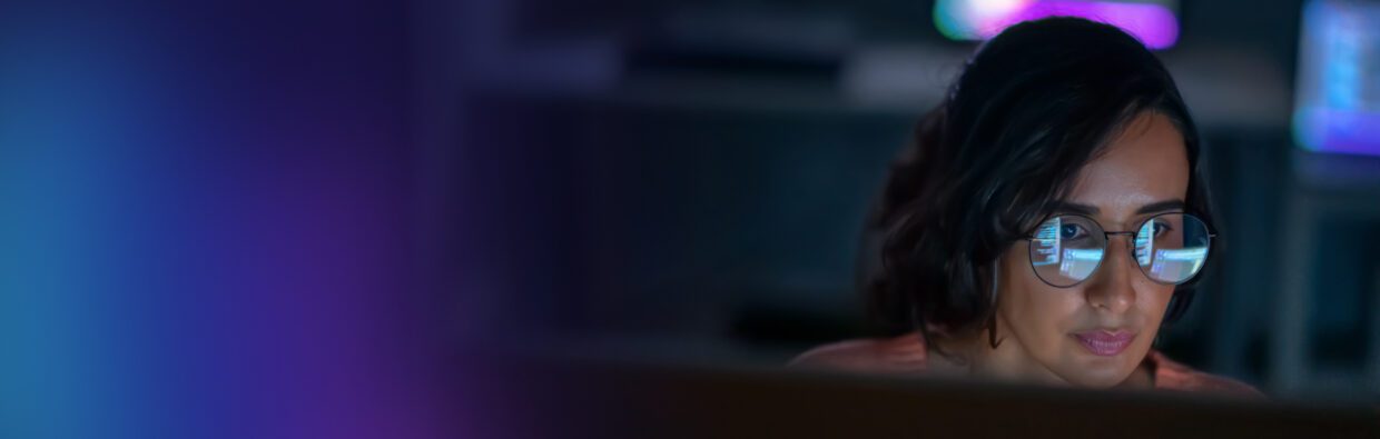 A woman wearing glasses focused on a computer screen in a dimly lit workspace, with colorful ambient lighting in the background, illustrating the concept of digital work. NIS2 explained.