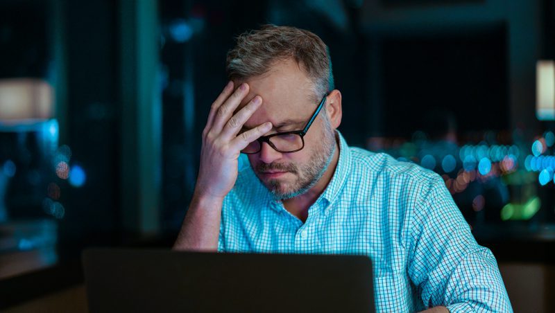 A man wearing glasses with short hair, seated at a desk in a dimly lit environment, looking stressed as he cradles his forehead while working on a laptop. He appears focused on the screen, indicating the task's complexity, relating to finding a managed security partner.