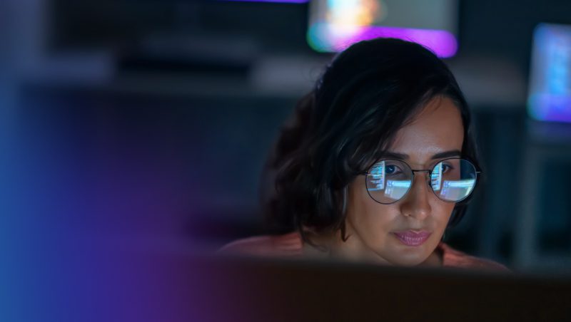 A woman wearing glasses working on a computer in a dimly lit environment, with colorful screens visible in the background. The focus is on her concentrated expression while examining data or information. NIS2 explained.