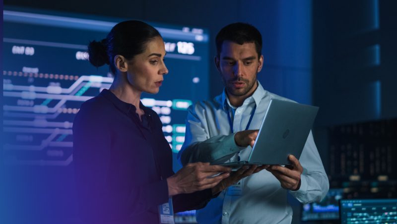 A woman and a man in a dark room, examining information on a laptop. The background features digital screens displaying data and graphs, indicating a tech-focused environment.