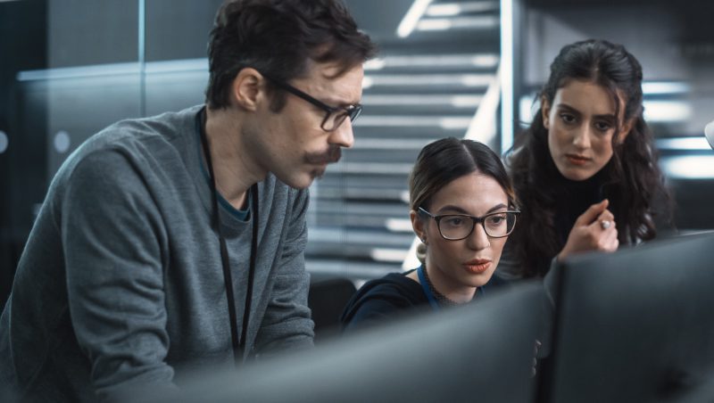 Three colleagues discussing data analysis in a high-tech office setting, with two women and one man focused on a computer screen, highlighting collaboration on 2023 security threats.