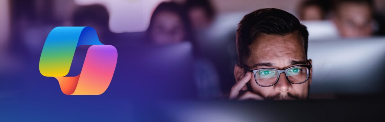 A man wearing glasses working intently in front of a computer screen, with blurred figures in the background. To the left, there is a colorful logo with a stylized design.