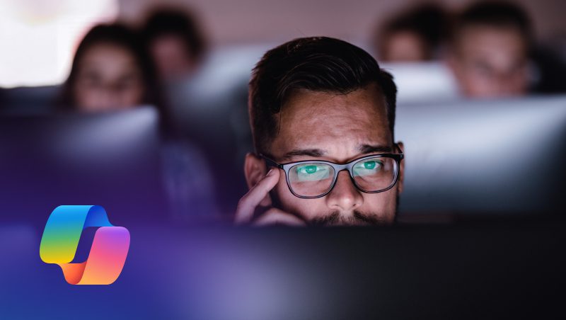 A man wearing glasses, looking intently at a computer screen in a dimly lit room, with blurred figures seated behind him, focused on their own screens. A colorful logo appears in the lower left corner.