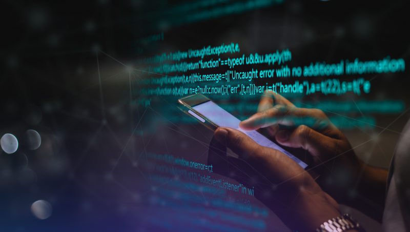 A person holding a smartphone while viewing programming code on a screen in the background. The code features error messages and programming syntax, creating a tech-focused atmosphere relevant to digital security and troubleshooting, including PlugX Utilising Steam.