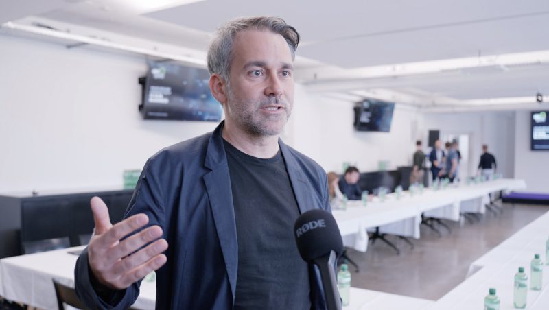 A man speaking into a microphone in a conference room. He is wearing a black jacket and black shirt. Tables are set up in the background with bottles of water. Screens are visible displaying presentations.