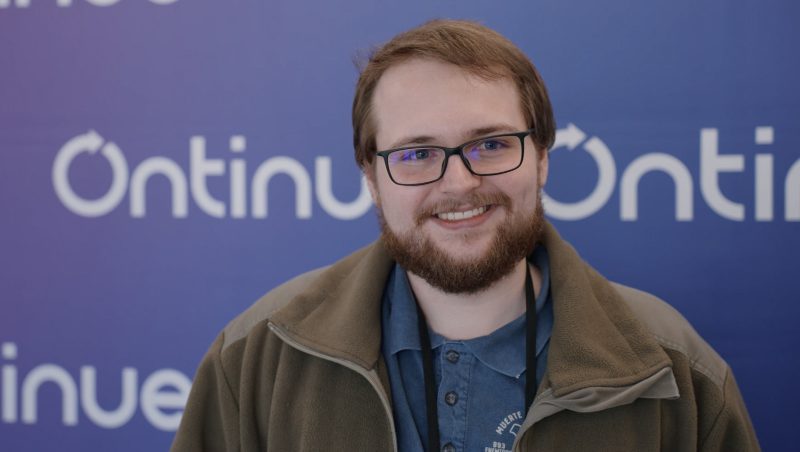 A smiling man with glasses and a beard wearing a green jacket and blue shirt, standing in front of a blue backdrop with the word 'Continue' visible.