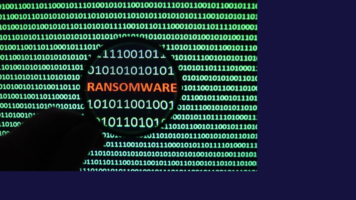 A close-up view of binary code on a digital screen, with the word 'RANSOMWARE' highlighted in orange through a magnifying glass. This image illustrates the concept of ransomware in digital security.