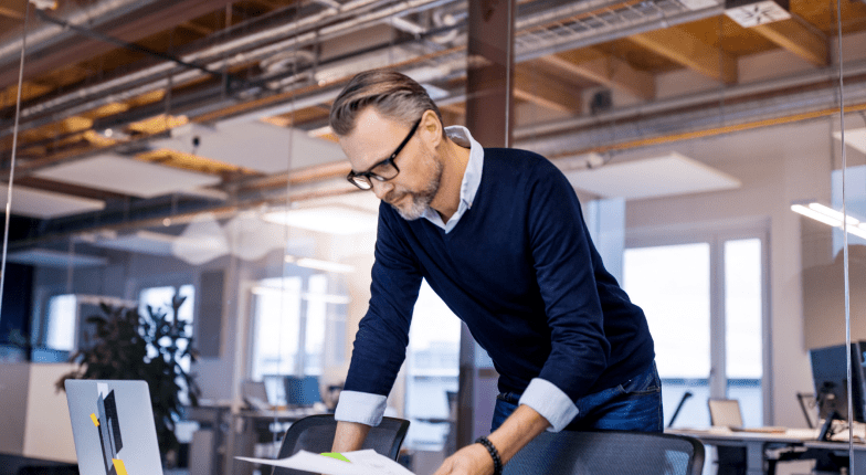 A man wearing glasses and a navy sweater is standing at a desk in a modern office, reviewing documents while surrounded by computer equipment.