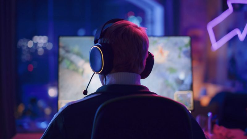 A person with short hair wearing a headset sits in front of a computer monitor displaying a video game. The room is illuminated with colorful LED lights, creating a gaming atmosphere.