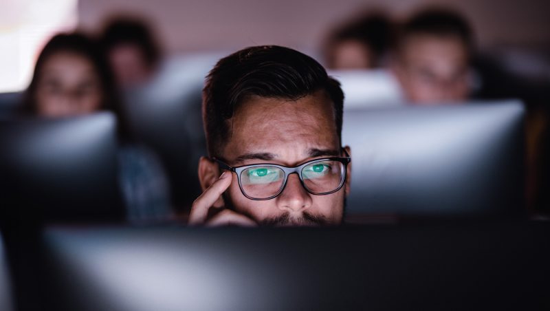 A man wearing glasses focused on a computer screen, with a contemplative expression. Several blurred figures are visible in the background, also engaged with their screens in a dimly lit environment.