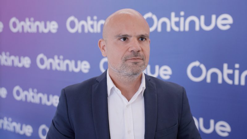 A man with a bald head and light beard wearing a dark suit and white shirt, standing in front of a blue backdrop with the logo 'Ontinue' in white.