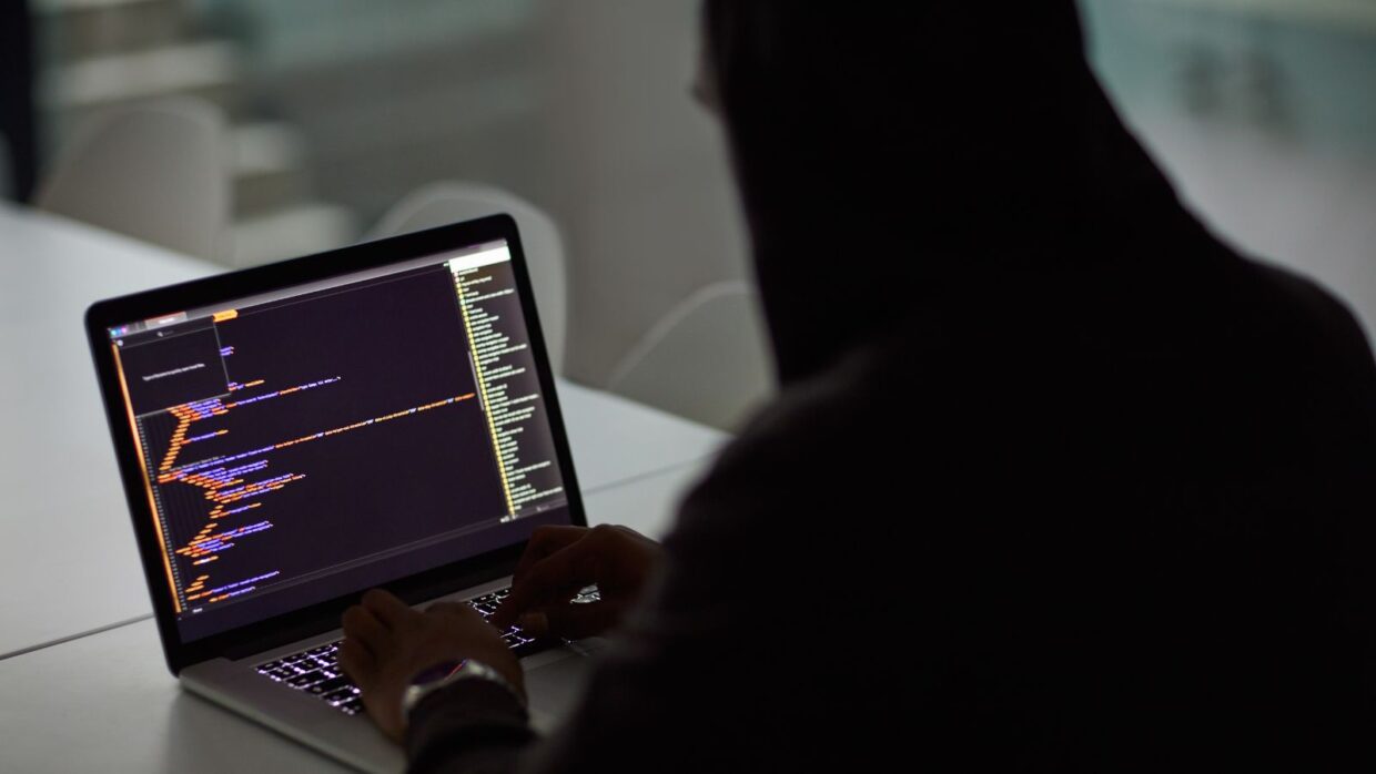 A person in a dark hoodie typing on a laptop displaying lines of code, with a soft focus background suggesting a tech environment. This image relates to the Rise of Infostealer Malware.