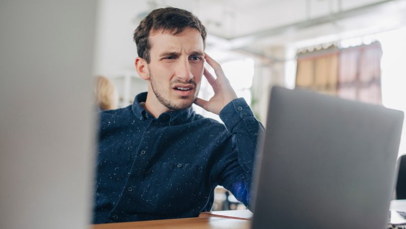 A man with confusion on his face, sitting at a desk and looking at a laptop screen in an office environment. Promoting the idea of Encouraging Employees to Report Security Issues.