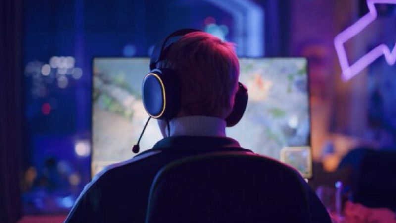 A person sitting in a gaming chair wearing a headset, focused on a computer monitor displaying a video game. The scene is illuminated by soft, colorful lighting from the window and room decor.