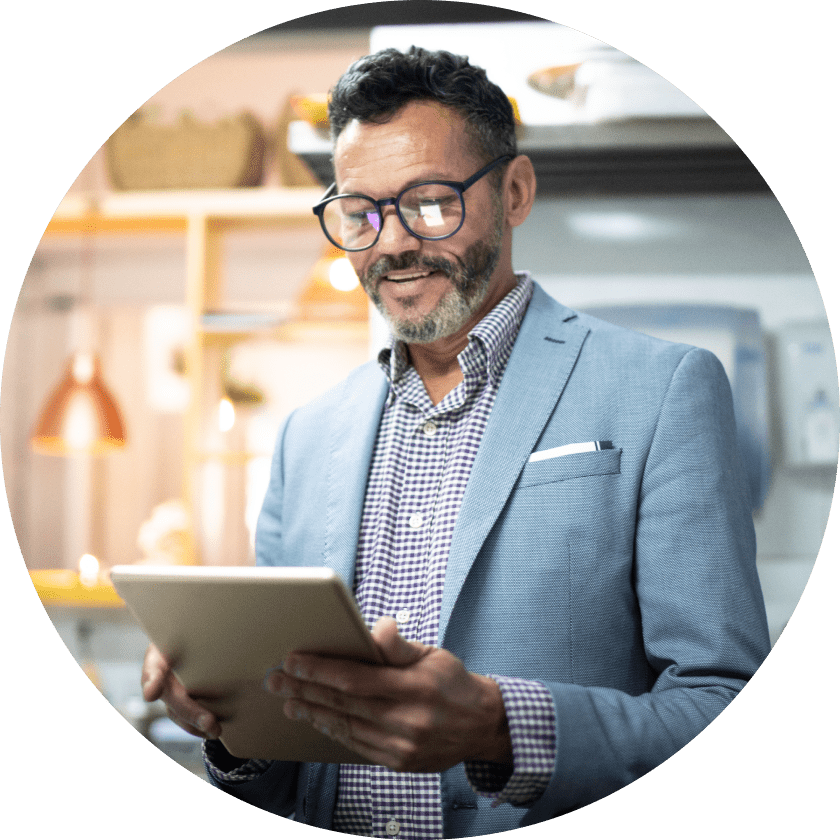 A middle-aged man with glasses smiles while holding a tablet in a well-lit kitchen setting, wearing a light blue blazer over a checked shirt.