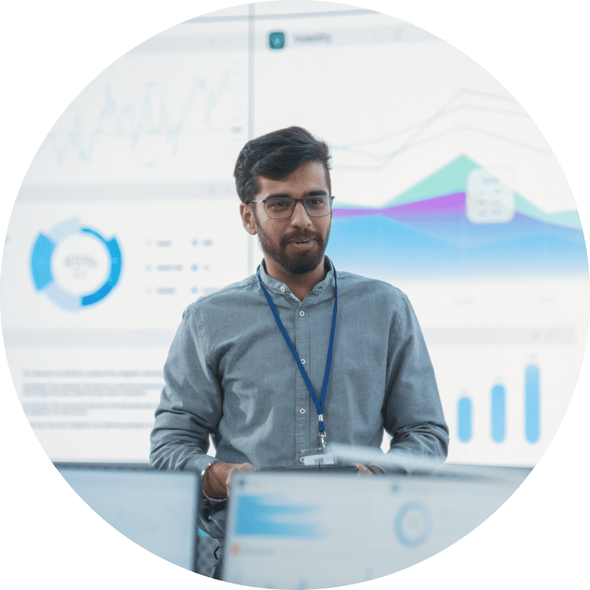 A man in a light blue shirt and glasses stands in front of a large screen displaying graphs and data visualizations in a modern office setting.
