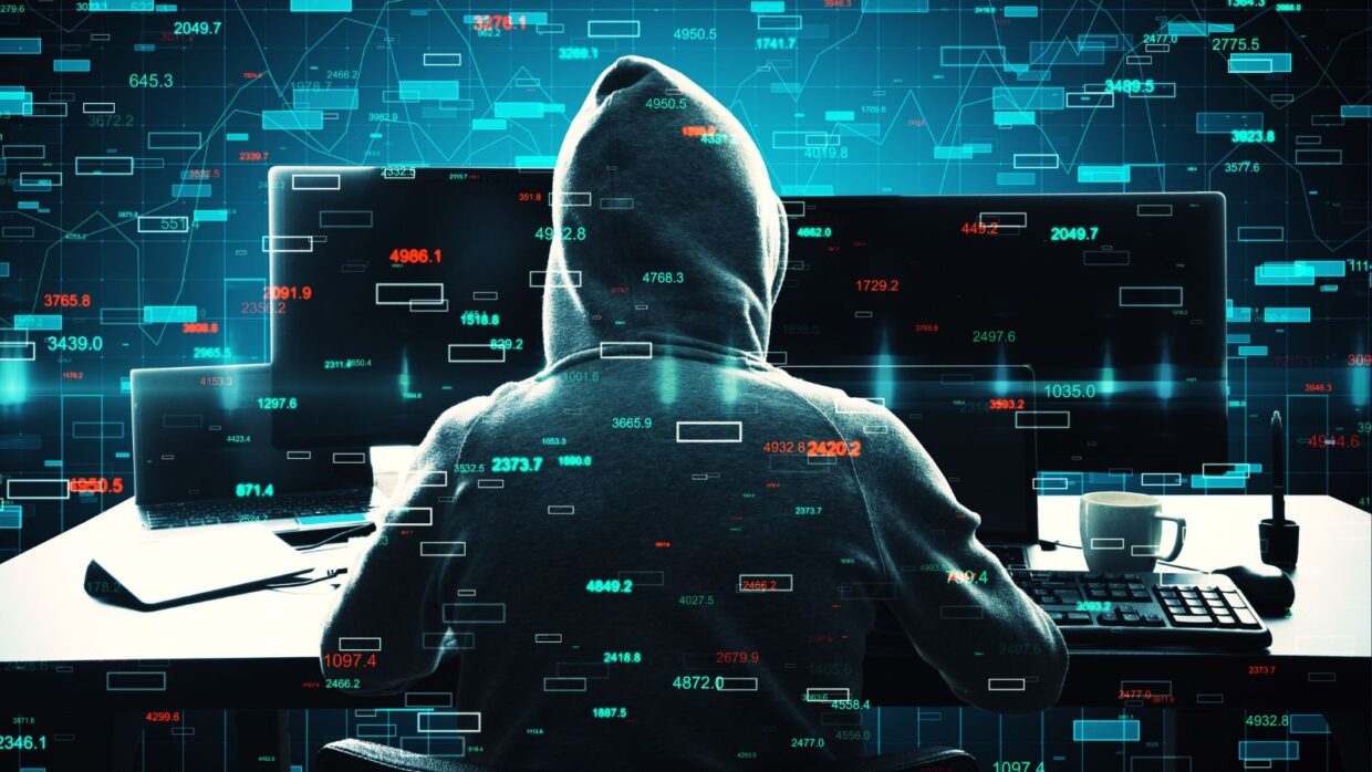 A person wearing a hoodie sitting at a computer with multiple screens displaying data and coding elements, illustrating the concept of detecting lumma infostealer.
