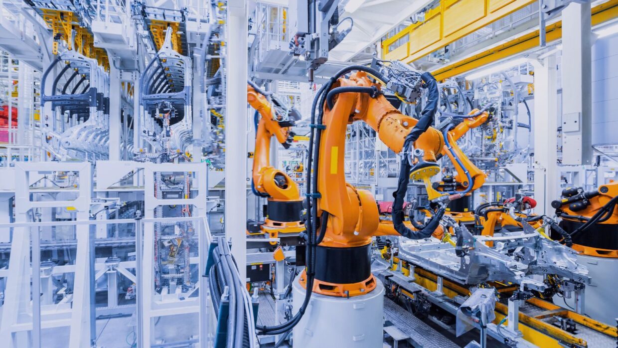 Industrial robots with orange arms working in a modern manufacturing facility, showcasing advanced automation technologies. The setup emphasizes Securing Operational Technology for efficient production.