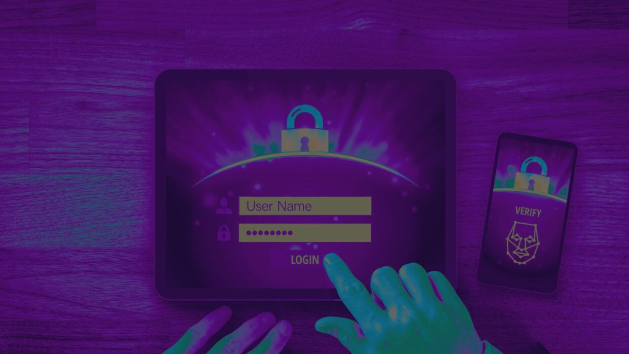 A person using a touchscreen tablet to enter login credentials, with a lock symbol on the screen. A smartphone displaying a verification prompt is nearby. This illustrates cyber criminals leveraging built-in Microsoft tools.