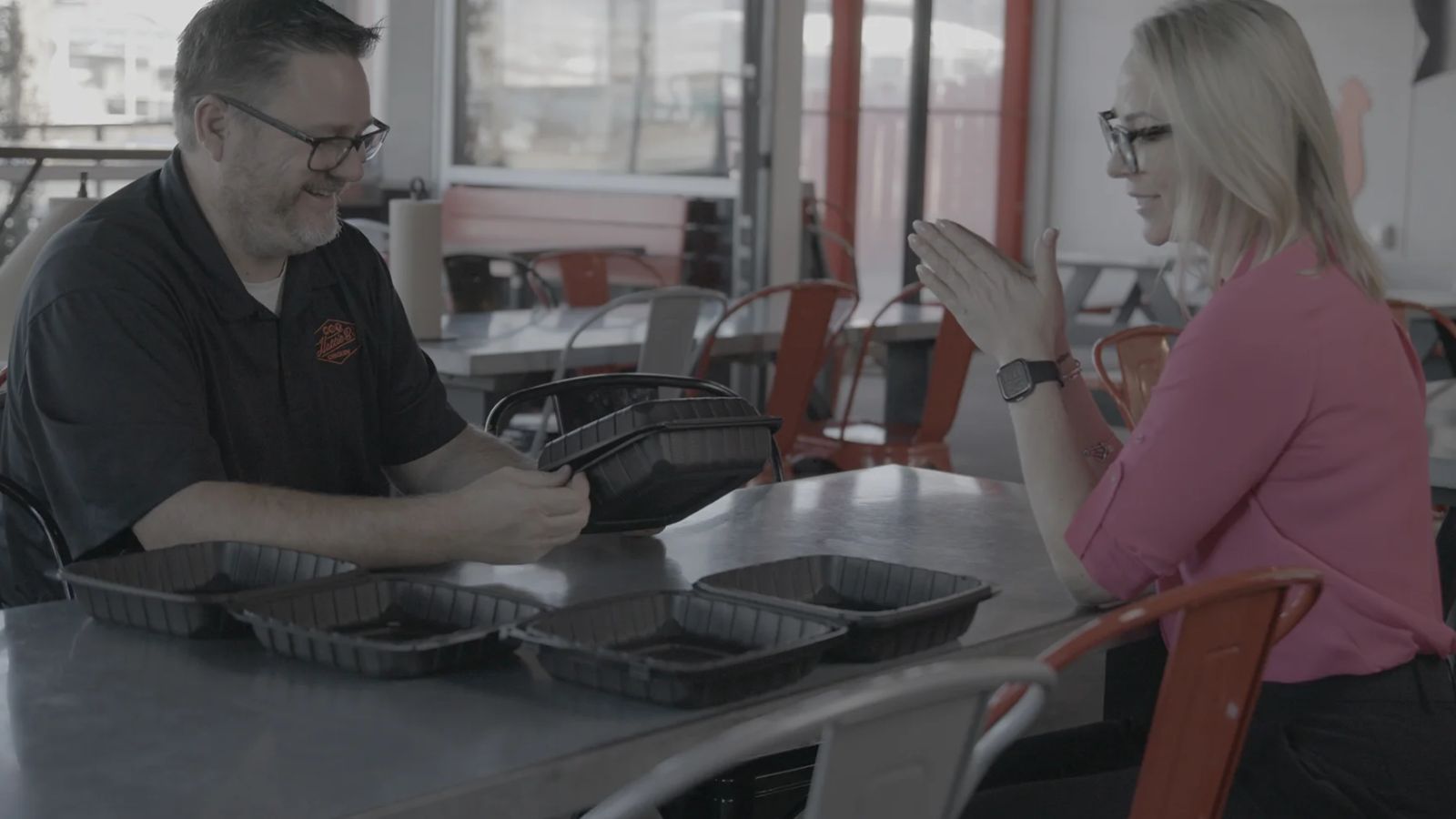 Two individuals seated at a table in a restaurant, with several black take-out containers in front of them. One person, wearing glasses and a black shirt, is examining the containers, while the other, in a pink shirt, is clapping her hands. This scene highlights teamwork and collaboration.
