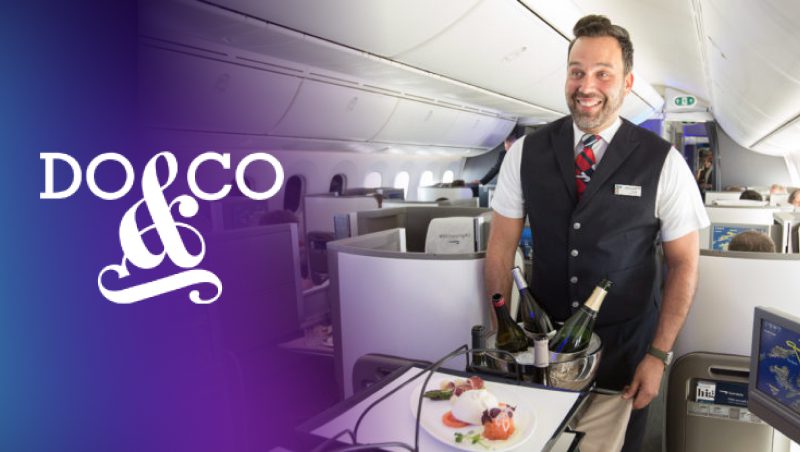 A flight attendant smiling while serving food and drinks in a modern airplane cabin, with a logo on the left side. The cabin features several rows of seats and large windows.