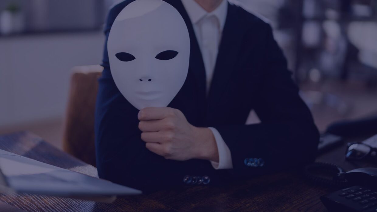 A person in a suit holding a white mask in front of their face, seated at a desk with office equipment around. The background is softly blurred, creating a focus on the figure and the mask, suggesting themes of identity or anonymity relevant to the Open AI June 2025 Report.