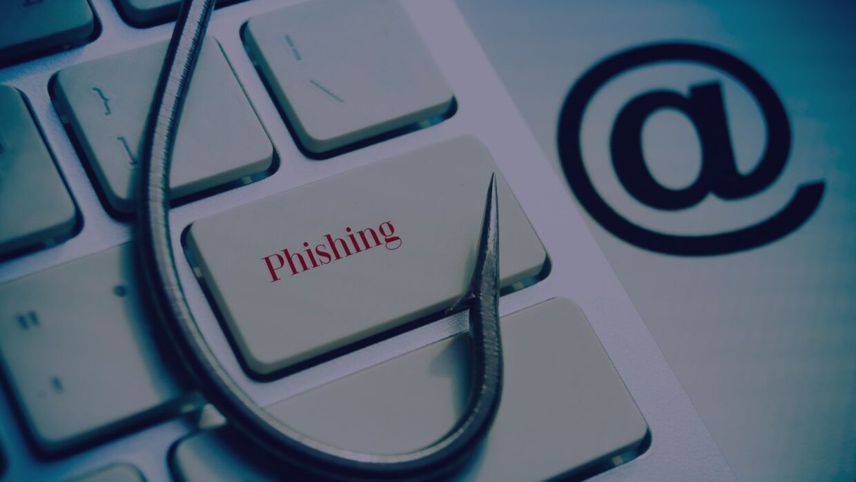 A close-up of a keyboard with a fishing hook resting on the 'phishing' key, alongside an '@' symbol in the background. This image illustrates the concept of Salty2FA Multi-Stage Phishing.