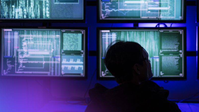 A person wearing a black hoodie sits in front of multiple computer monitors displaying code and data analysis, illuminated by blue and green lights in a dark environment.