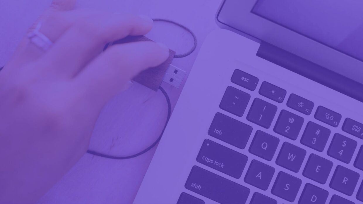 A hand holding a USB flash drive over a laptop's USB port, with the laptop keyboard visible. The image is tinted purple, emphasizing the action of connecting the USB-borne malware to the device.