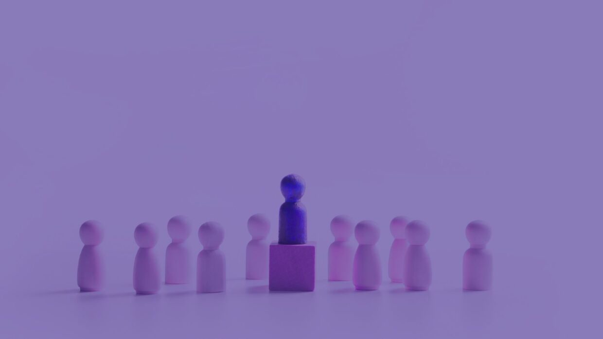 A group of pink wooden figurines surrounds a single blue figurine positioned on a purple block, illustrating a theme of leadership and collaboration, relevant to "Building Trust with AI".