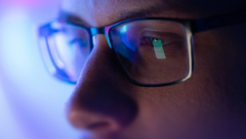 A close-up of a person wearing black rectangular glasses, with blue screen reflections on the lenses and a blurred face in the background.